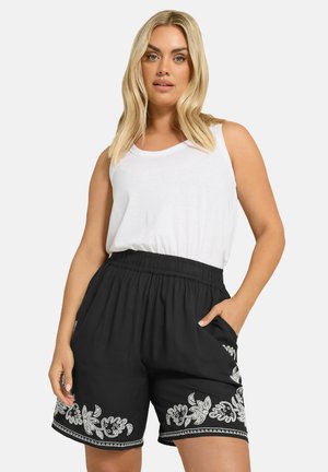 Pantaloni neri con ricami floreali bianchi all'orlo, dotati di una cintura elastica e tasche laterali, abbinati a una canotta bianca semplice.