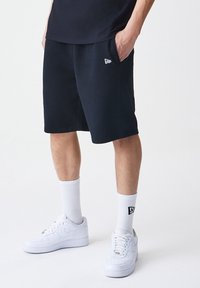 Sorte idrettsshorts laget av bomullsblanding, med snor i livet, sidelommer og liten logo på venstre side. Brukt med hvite joggesko og sokker.