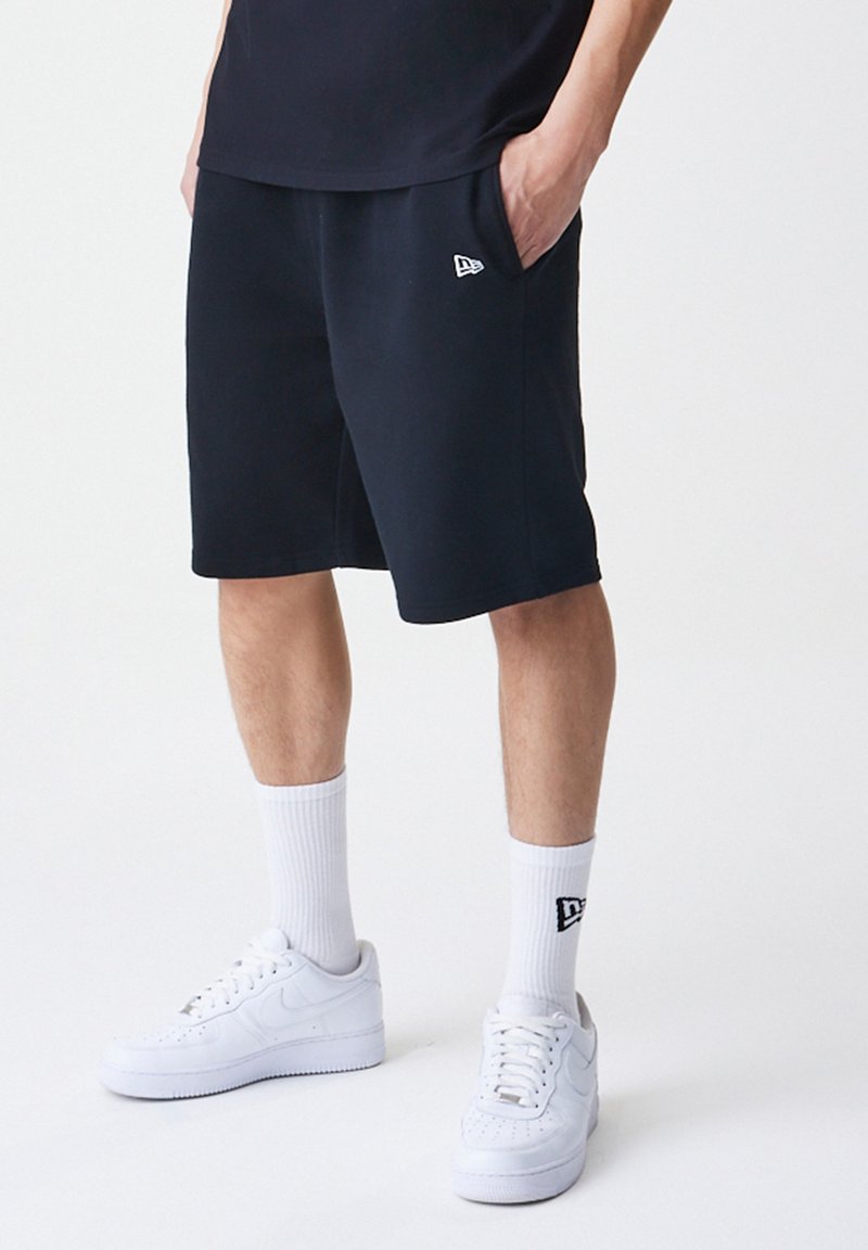 Sorte idrettsshorts laget av bomullsblanding, med snor i livet, sidelommer og liten logo på venstre side. Brukt med hvite joggesko og sokker.