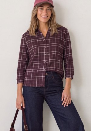 Jeune femme portant une chemise bordeaux à carreaux boutonnée, un jean foncé et une casquette assortie, tenant un sac à main bordeaux contre un mur uni.