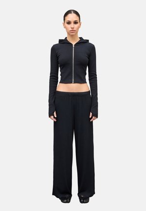 Donna in piedi che indossa una felpa nera corta con zip e foro per il pollice, pantaloni neri a gamba larga e scarpe nere su uno sfondo chiaro uniforme.