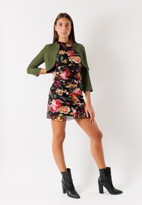 Abito floreale nero, con vivaci motivi rosa e gialli, abbinato a un giubbotto verde corto. Stivali neri alla caviglia completano il look.