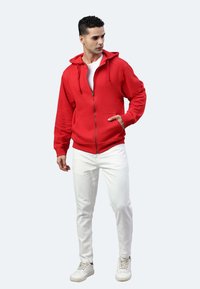 Felpa rossa con zip, cappuccio regolabile, tasca a marsupio e polsini a costine, abbinata a pantaloni bianchi e scarpe da ginnastica bianche.
