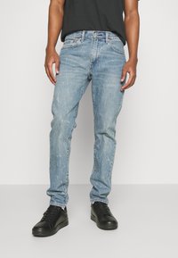 Vaqueros de mezclilla azul claro con corte recto, textura desgastada y detalles de desvanecimiento sutil, usados con zapatillas negras de corte bajo.