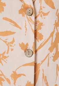 Beige tyg med orangea blommönster; har två runda knappar med en strukturerad yta; slät textur och lätt design.