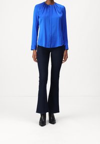 Kvinna som bär en kungligt blå långärmad blus, mörka utsvängda jeans och svarta klackade ankelskor stående mot en vit bakgrund.