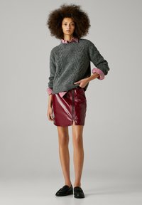 Un pull en maille texturée gris superposé sur une chemise à rayures rouges et blanches, associé à une jupe en faux cuir bordeaux brillant avec un détail de fermeture éclair.