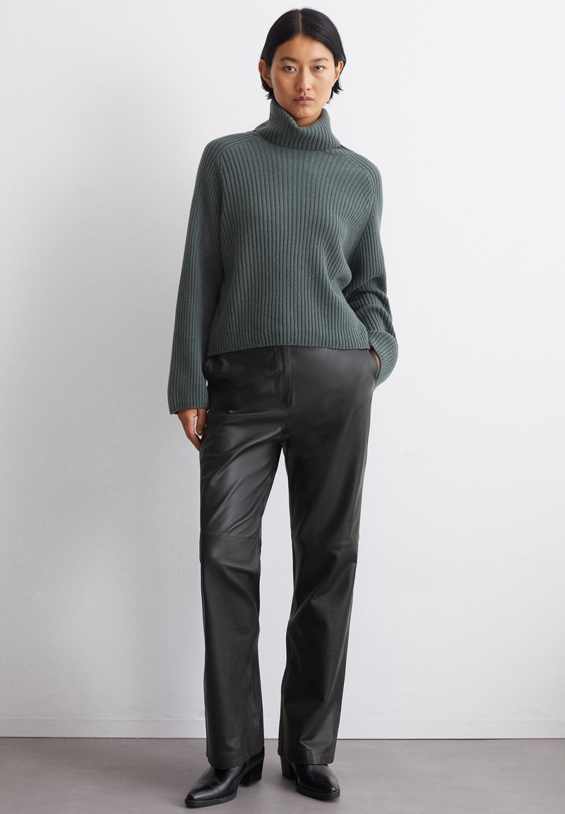 Grüner gerippter Rollkragenpullover kombiniert mit schwarzen Leder-Hose im geraden Schnitt und schwarzen Absatzstiefeln. Schlichtes, strukturiertes Design mit subtiler Textur.
