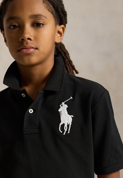 Laps punotulla tukalla, musta poolopaidan, jossa valkoinen kirjailtu poolopelaajan logo rinnassa, kaulus pystyssä.