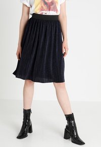 Falda plisada azul marino con una cinturilla elástica negra. Modelo combinada con botas de tacón negro brillantes y una camiseta gráfica.
