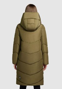 Person med lång olivgrön vadderad vinterjacka med stor quiltad huva, visad bakifrån mot en enkel ljus bakgrund.