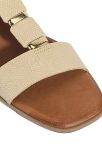 Beige gestructureerde leren sandaal met brede band en gouden hardware, geplaatst op een bruin leren voetbed. Rond neusontwerp.