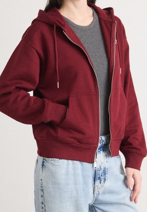 Personne portant un sweat à capuche zippé bordeaux sur un t-shirt gris et un jean bleu clair délavé à l'acide, une main dans la poche du sweat.