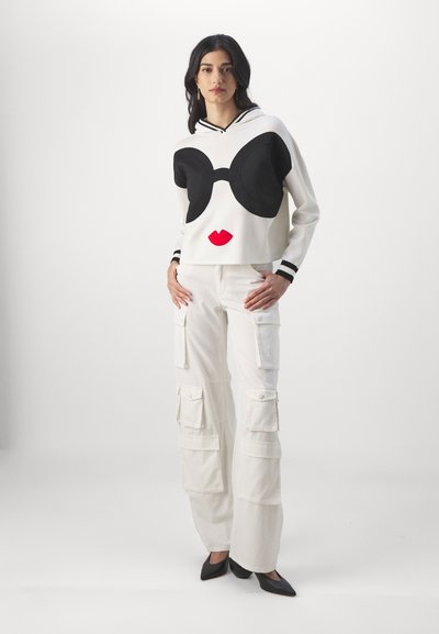 Pull court blanc avec un grand motif de lunettes de soleil noires et de lèvres rouges. Associé à un pantalon cargo blanc et des ballerines noires à bout pointu.