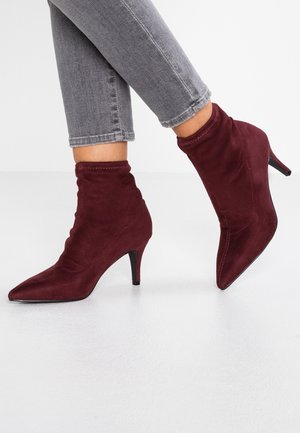 Personne portant un jean gris ajusté et des bottines en daim bordeaux à bout pointu avec talons petits sur un sol blanc.