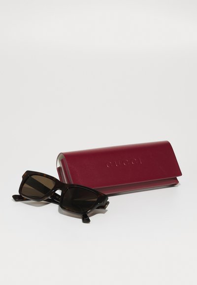 Barna teknősbéka napszemüveg sötét lencsékkel, egy bordó bőr tok mellett pihen, amelyen a "GUCCI" felirat látható. A tok elegáns formatervezéssel rendelkezik.