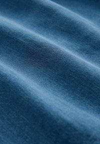 Plavi traper tkanina s suptilnom teksturom i blagim sjajem; ima glatke, drapirane nabore koji naglašavaju mekoću materijala.