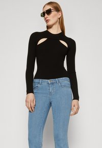 Bodysuit negro de mangas largas con detalles de corte, combinado con jeans de denim azul claro. La modelo lleva gafas de sol oscuras y está de pie contra un fondo gris.