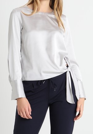 Silberne Satinbluse mit langen Ärmeln, rundem Ausschnitt und seitlichem Bindeband. Sie zeichnet sich durch eine glatte Textur und einen leicht lockeren Schnitt aus.