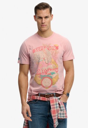 Uomo che indossa una maglietta rosa con design ispirato al Giappone, jeans blu, camicia a quadri annodata in vita e orologio da polso.