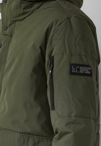 Doudoune vert olive avec une poche zippée sur la manche gauche et un patch de marque noir. Caractérisée par un tissu texturé et une fermeture à pression.