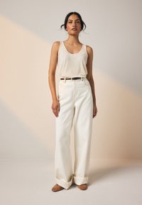 Pantalon blanc à jambes larges, associé à un débardeur côtelé beige clair, souligné par une ceinture noire et des chaussures marron. Tissu lisse, coupe décontractée.