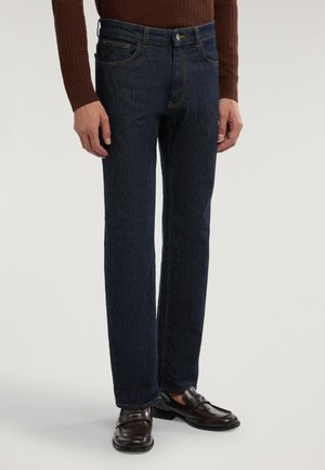 Man klädd i mörkblå raka jeans, brun tröja och mörkbruna läderloafers, står mot en enfärgad ljus bakgrund.
