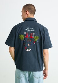 Kortermet marineblå skjorte med fargerik brodert scene på baksiden med figurer, trær og høyttalere; avslappet passform med en uformell stil.