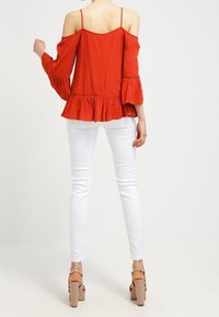 Haut rouge hors-épaule avec des manches évasées et un volant à l'avant, associé à un jean skinny blanc et des sandales à talons beiges.