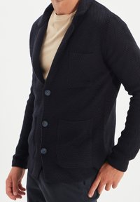 Cárdigan texturizado azul marino con cuello chal, tres botones y dos bolsillos delanteros. Hecho de una tela tejida suave para mayor comodidad.