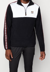 Svart fleece-pullover med vit övre panel, dragkedja vid kragen och röda accenter. Har logotyp på bröstet och mönstrade sidostickningar. Standard passform.