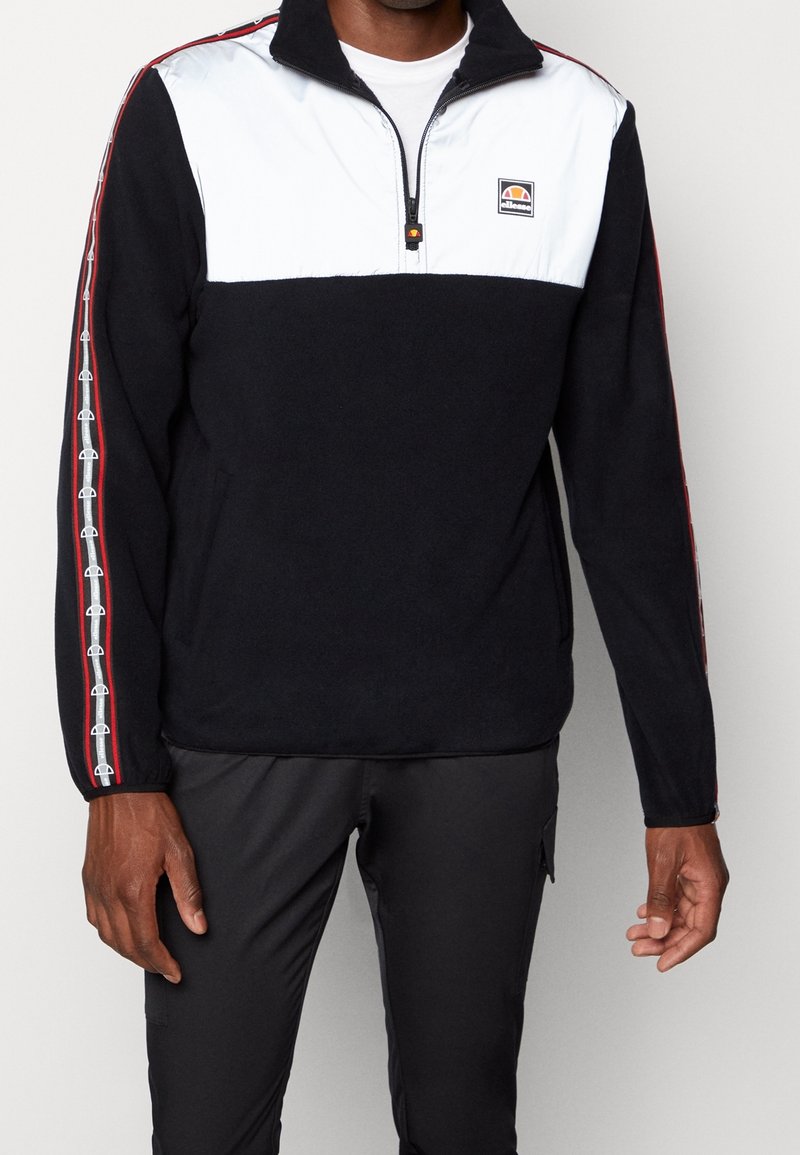 Svart fleece-pullover med vit övre panel, dragkedja vid kragen och röda accenter. Har logotyp på bröstet och mönstrade sidostickningar. Standard passform.