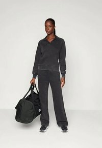 Conjunto de pantalones de chándal y sudadera negra con cuello con cremallera, hecho de una tela suave. El modelo sostiene un gran bolso de deporte negro y lleva zapatillas negras.