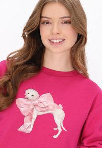 Ragyogó rózsaszín pulóver fehér kutyás grafikával, amelyet egy nagy rózsaszín masni díszít. Puha anyagú és kényelmes, laza szabású.