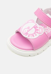 Růžové sandály se hladkým syntetickým svrškem, s grafikou medvěda a bílým páskem. Tlustá bílá podrážka s texturovaným vzorem.