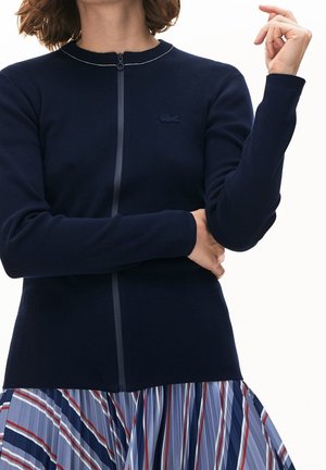 Vrouw draagt een marineblauwe trui met rits en een klein logo, en een geplooide rok met blauwe, witte en rode strepen, armen gekruist en opgeheven.