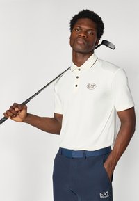 Ανδρικό πόλο γκολφ κοντό μανίκι, ανοιχτό κρέμα με σκούρα κουμπιά, λογότυπο στο στήθος, συνδυασμένο με ναυτικές παντελόνες και πλεκτή μπλε ζώνη.