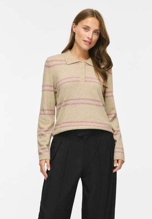 Femme aux longs cheveux bruns portant un polo à manches longues beige et rose à rayures et un pantalon large plissé noir, debout sur fond blanc.