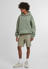 Green hoodie with a front pocket and white logo, paired with khaki cargo shorts and light sneakers. Model has curly hair and white socks.