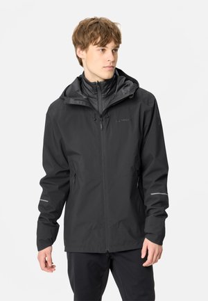 Jeune homme portant une veste imperméable noire à capuche avec des poches zippées, regardant sur le côté sur un fond clair uni.