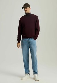 Mężczyzna w ciemno-burgundowym golfie, jasnoniebieskich dżinsach, białych sneakersach i ciemno-brązowej czapce bejsbolowej, stojący na tle jednolitego tła.