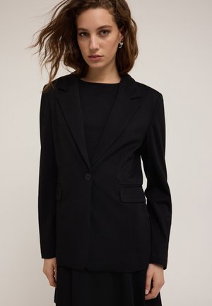 Blazer nero su misura realizzato in tessuto morbido, dotato di chiusura con un solo bottone, rever a punta e due tasche frontali. Vestibilità strutturata.