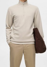 Sweat à zip beige avec un col montant, en tissu doux. Assorti à un pantalon kaki clair et tenant une veste marron.