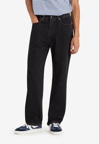 Homme portant un jean noir ample, une chemise rayée bleue et blanche, et des baskets bleu et blanc, debout avec une main dans la poche.