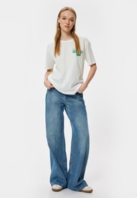 Hvid bomulds T-shirt med korte ærmer, grøn trykt tekst; parret med lyseblå brede denim jeans og beige sneakers. Simpelt, afslappet design.