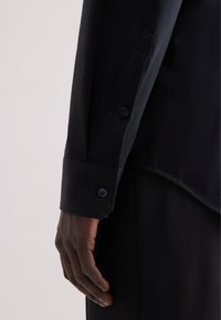Camicia nera a maniche lunghe con polsini abbottonati, marchio discreto sulla manica e una texture di tessuto liscia.