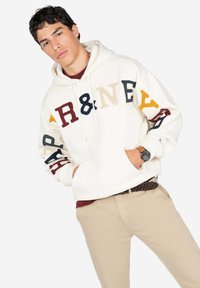Sudadera color crema con letras bordadas multicolores. Presenta una capucha con cordón y puños acanalados. Combinada con pantalones beige.