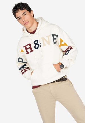 Sudadera color crema con letras bordadas multicolores. Presenta una capucha con cordón y puños acanalados. Combinada con pantalones beige.
