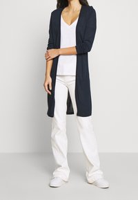 Cardigan long marine avec une ouverture devant et des manches longues, associé à un t-shirt blanc à col en V et à un pantalon blanc, présentant une silhouette décontractée.