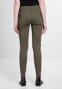 Persona, ģērbjoties olīvu zaļās šaurās biksēs un melnās potīšu zābakos ar aizmugurējām rāvējslēdzēm, stāv pretī neitrālai gaišai fona.